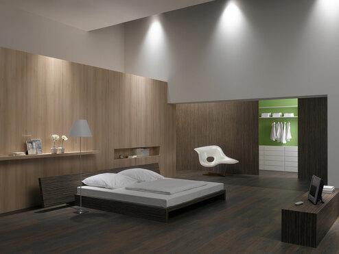 Schlafzimmerimpression von Bett, Nachtisch und Sideboard mit Holzverkleidung an Wand und indirektert Beleuchtung